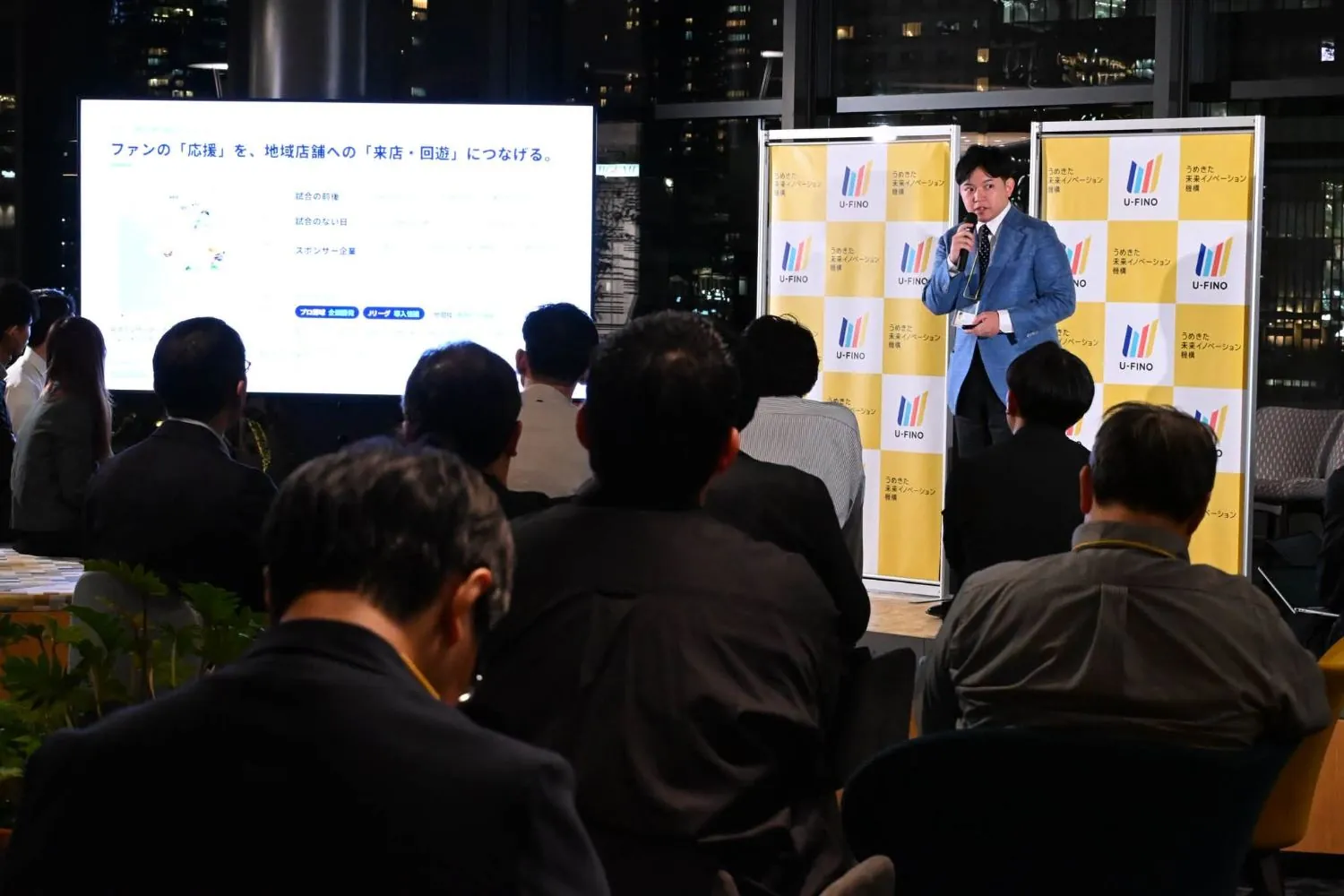 ステージで、スポーツのファンの応援を地域店舗への来店・回遊につなげる取り組みを説明する様子